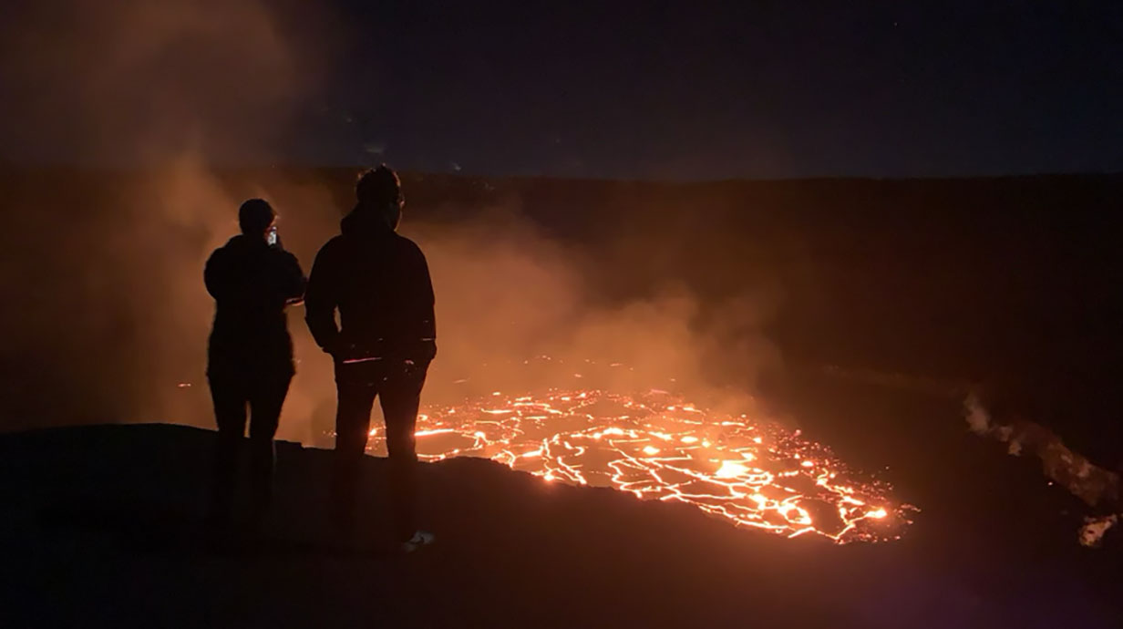 Silhouette of two people at night standing on a lookout and looking at lava in a volcano from below.