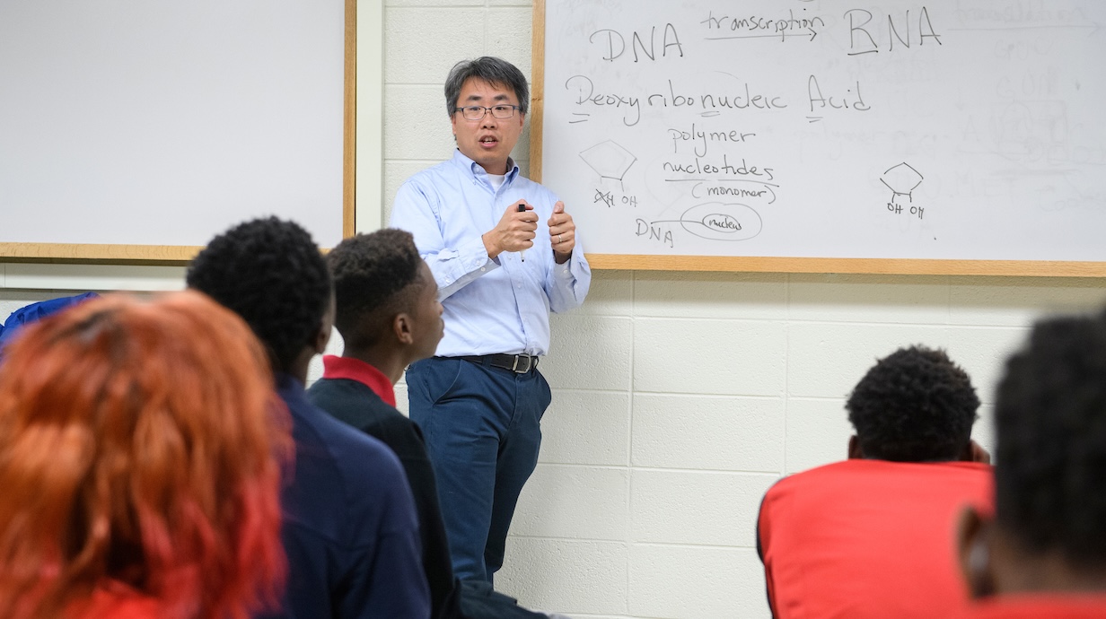 male professor standing in front of a white board while teaching a class full of students