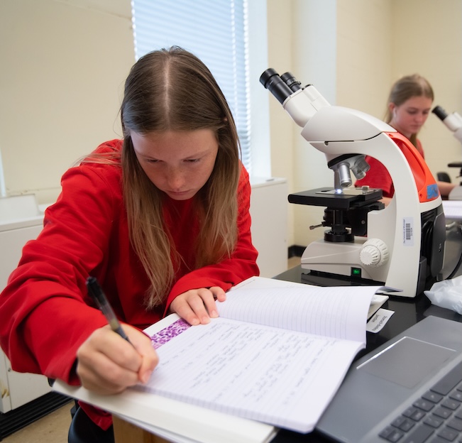 two girls sit at a table with a microscope on the table in front of them and take notes