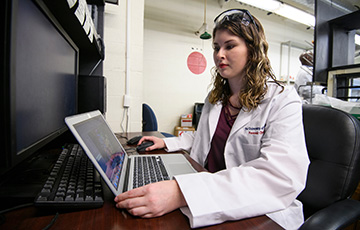 female student in white lab coat working at computer