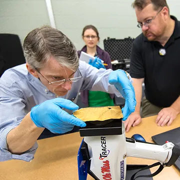 Researcher wearing glasses and blue vinyl gloves bent over machine holding sample. Two onlookers in background.