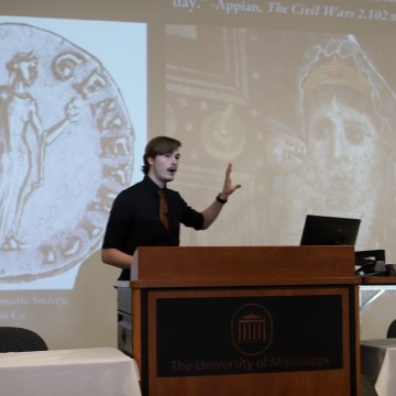 student standing at a lectern while making a presentation with images of ancient artifacts in the background