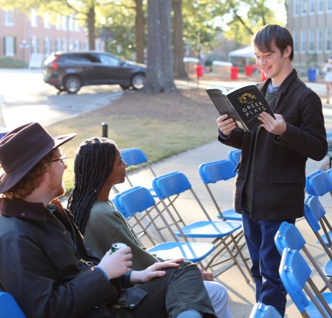 student holding a book and reading a Greek play outloud to a small audience while standing outside