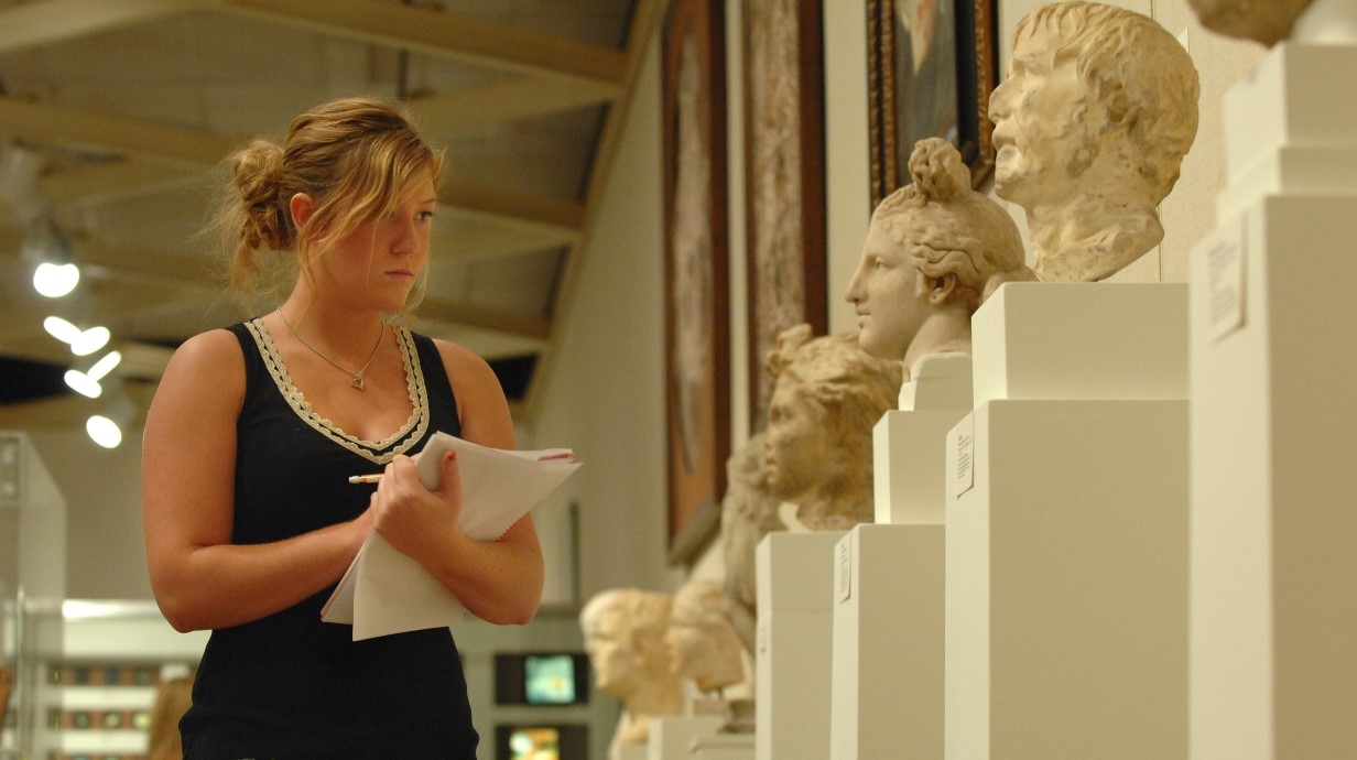 Student holding papers as she looks at ancient Greek artifacts of statue busts in museum