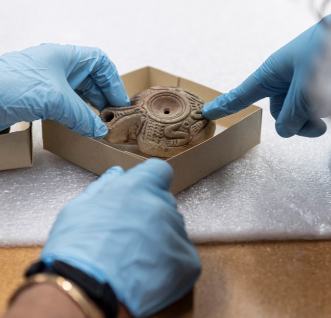 image of ancient clay object from the UM Museum connection with gloved hands and touching and pointing to it