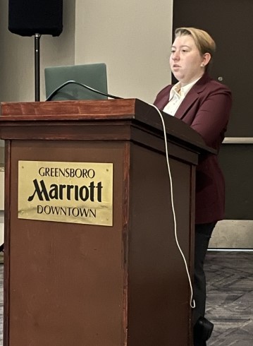 image of Gwen Pfrenger standing and talking at a lectern with the plaque of the Marriott hotel