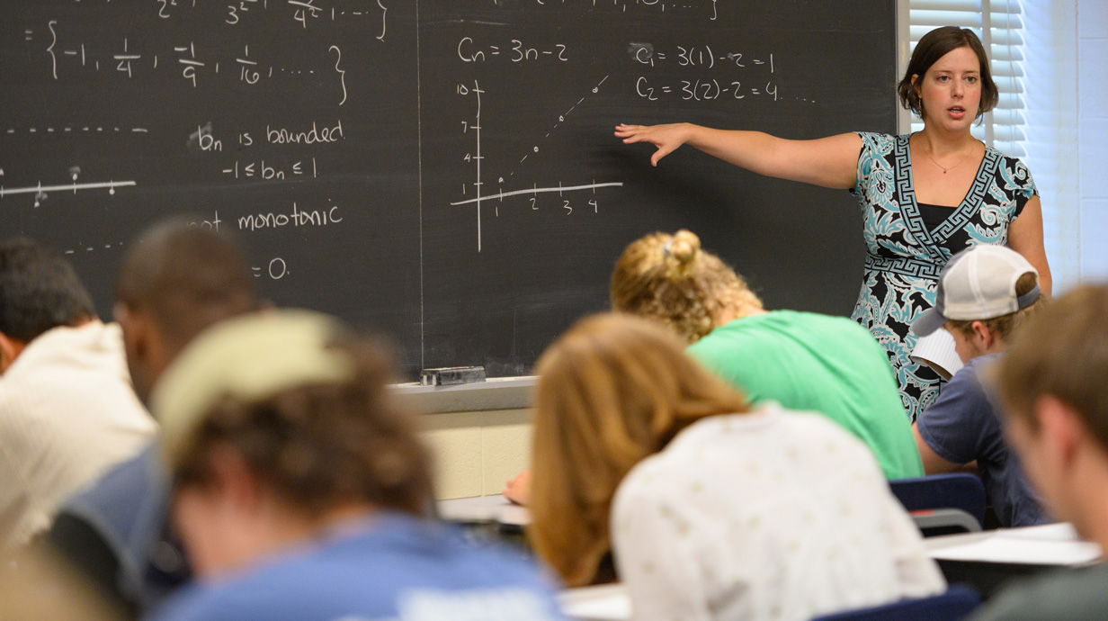 female professor pointing to a whiteboard and teaching in front of a class