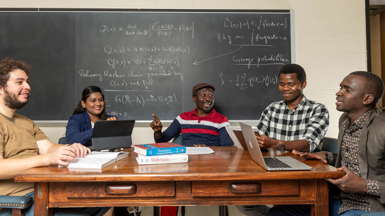 professor and students sit around table and discuss math with a chalk board in the background