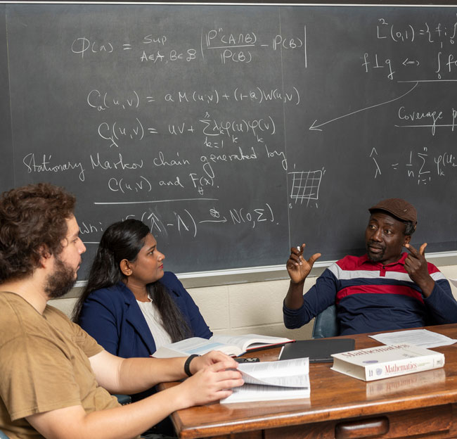 male professor sitting across from two students with math functions on a chalk board in the background