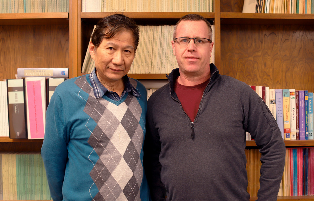 Two male professors stand in front of a bookcase and look into the camera.