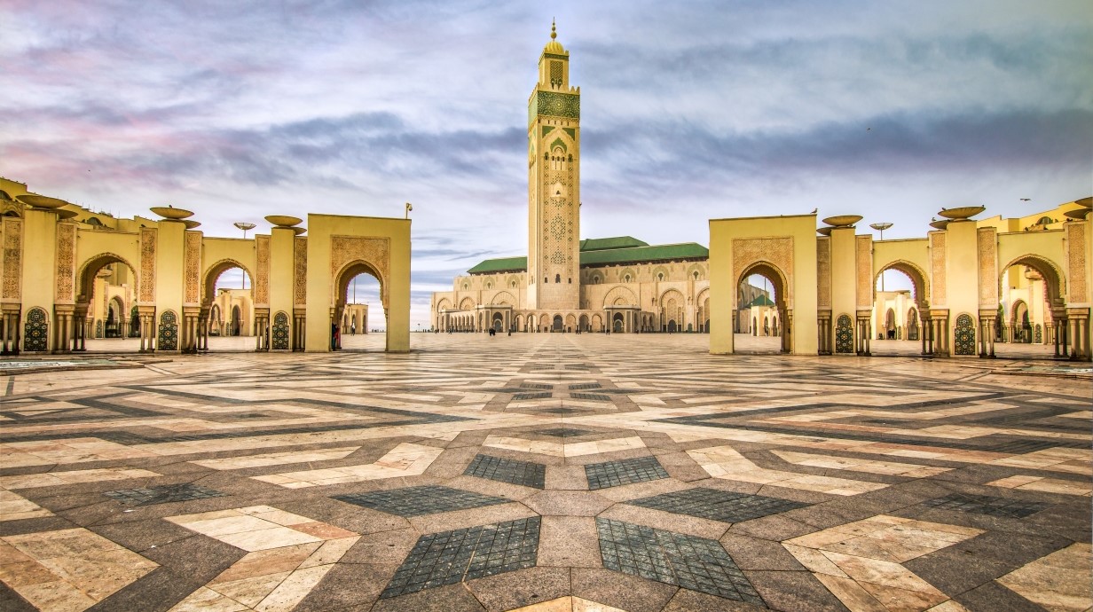 image of a large mosque in Morocco. It has a large marble tile square with designs in the tile and the mosque building has arched doorways and a large tower