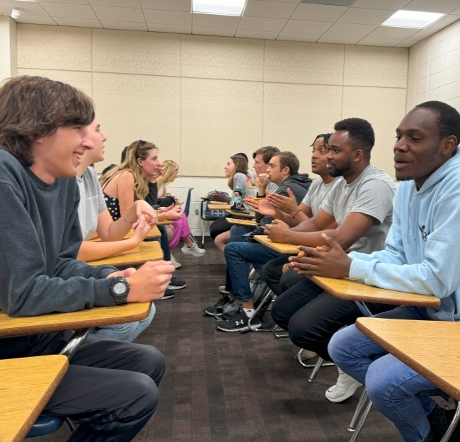 a group of students seated in two rows facing each other as they converse in French in a classroom 