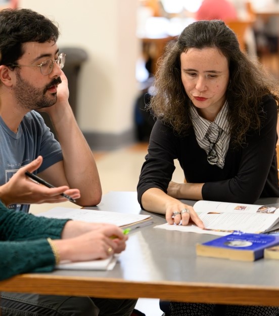 student and faculty member sitting together at a table and talking about a book in front of them. 