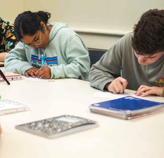 image of students seated at a table looking down at the papers in front of them as they write