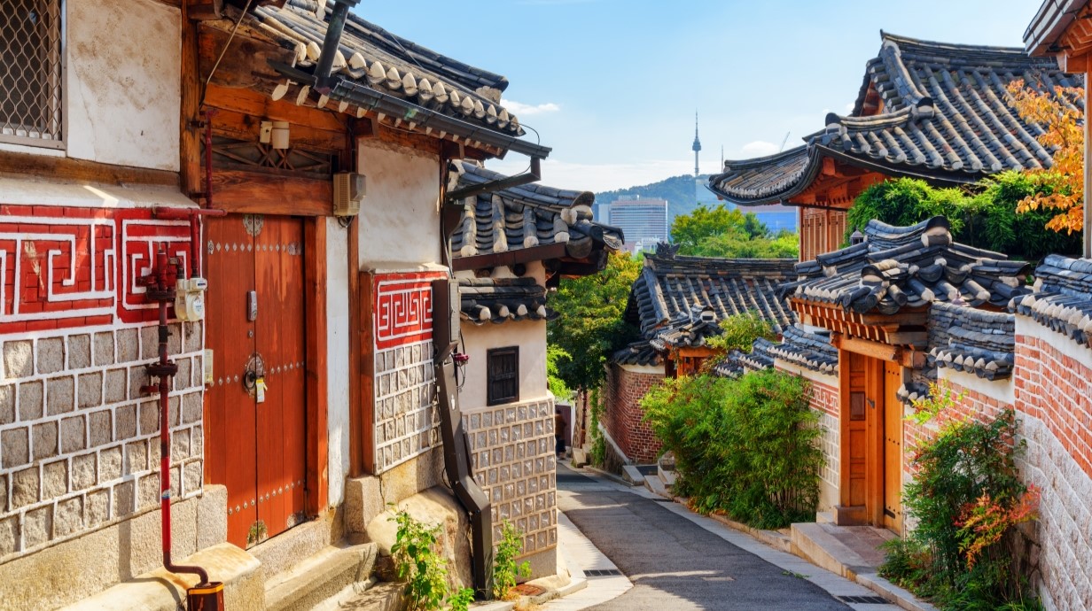 View of cozy old narrow street and traditional Korean houses of Bukchon Hanok Village in Seoul, South Korea. Seoul Tower on Namsan Mountain is visible on blue sky background. Scenic cityscape.