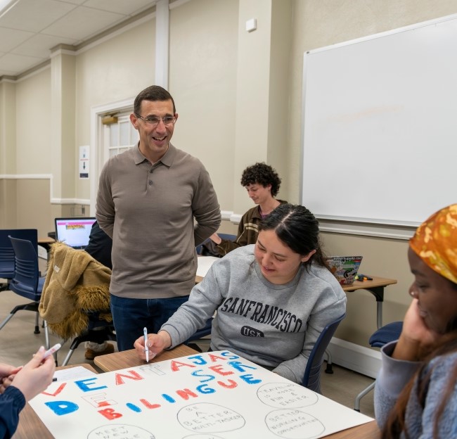 image of Dr. Fafulas standing in a classroom and looking on as students work on a poster board 