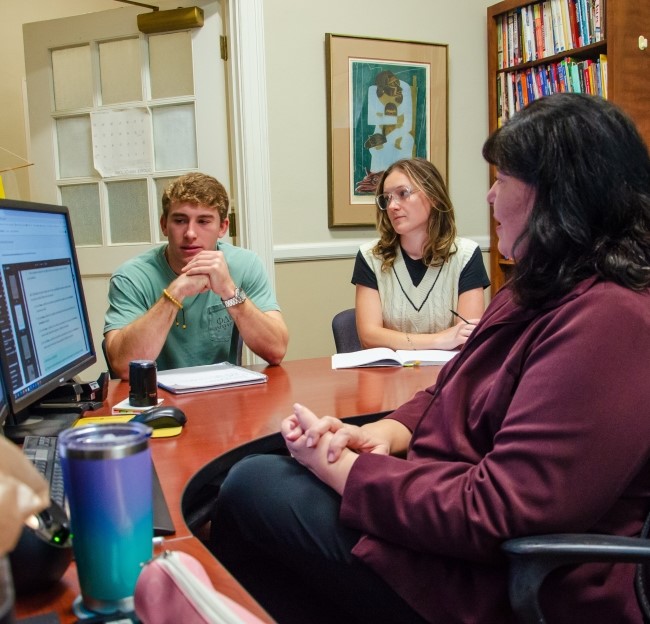 image of professor and two students seated around the desk in a professor's office. they are talking and looking at text on a computer screen