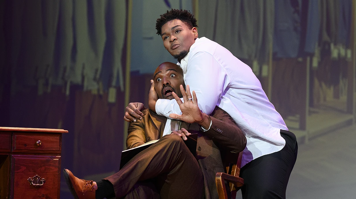 A performer during The Light in the Piazza wraps his arms around the head of another performer sitting in a chair as he raises his hand up with a shocked look on his face