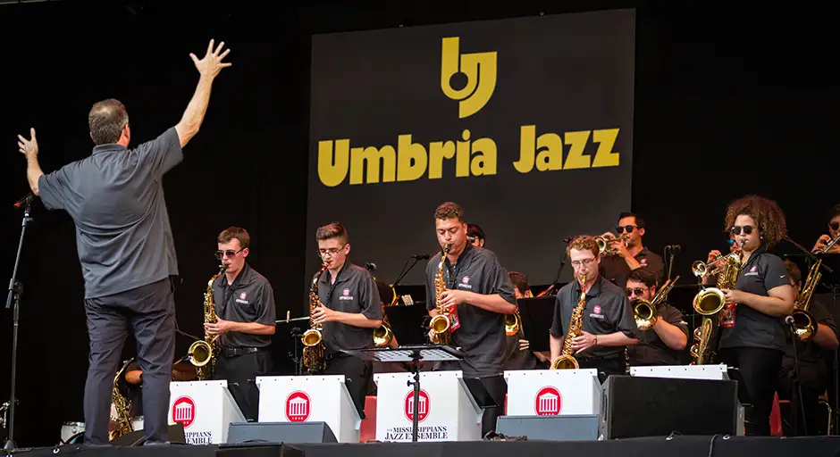 Jazz musicians and conductor performing a concert on stage with "Umbria Jazz" on banner behind them.