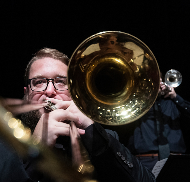 trombone player wearing a suit and glasses stares straight at the camera while playing his instrument