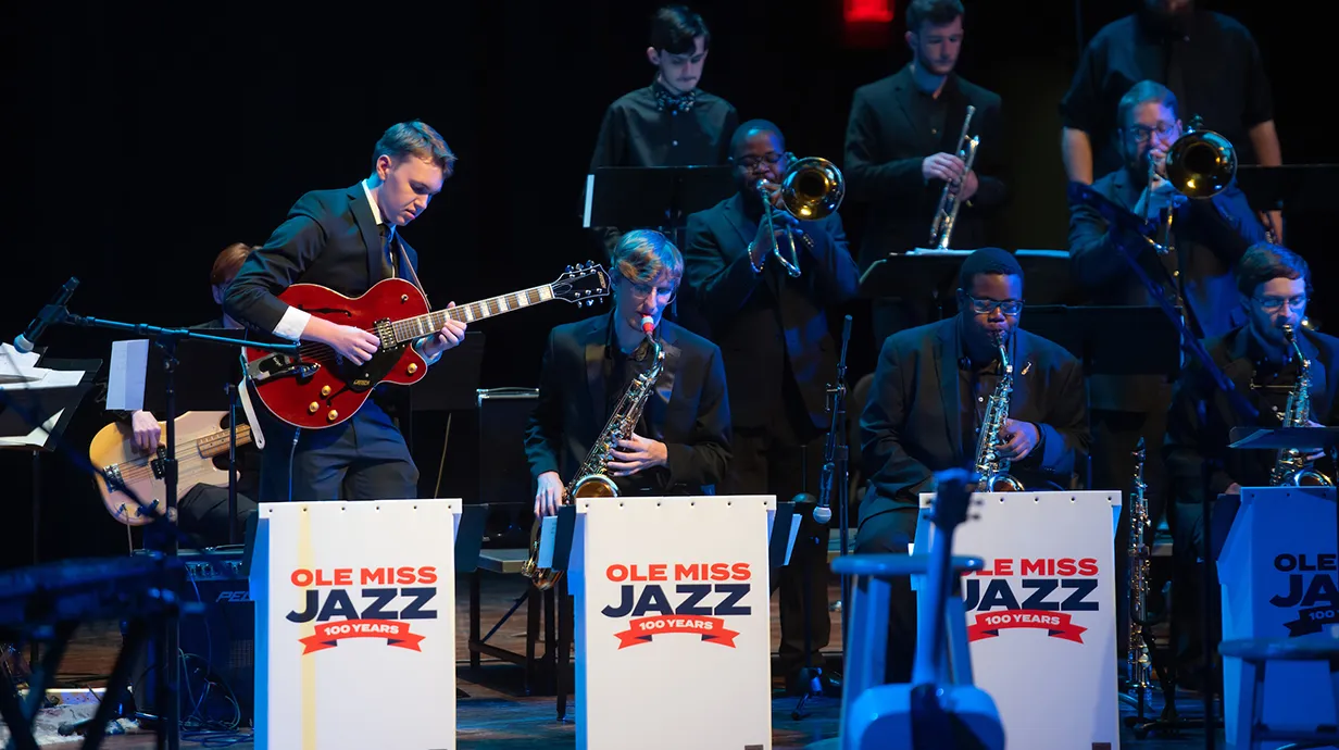 Stage with jazz band members. Man on left playing guitar; three other musicians playing a saxophone. Banners read "Ole Miss Jazz 100 Years"