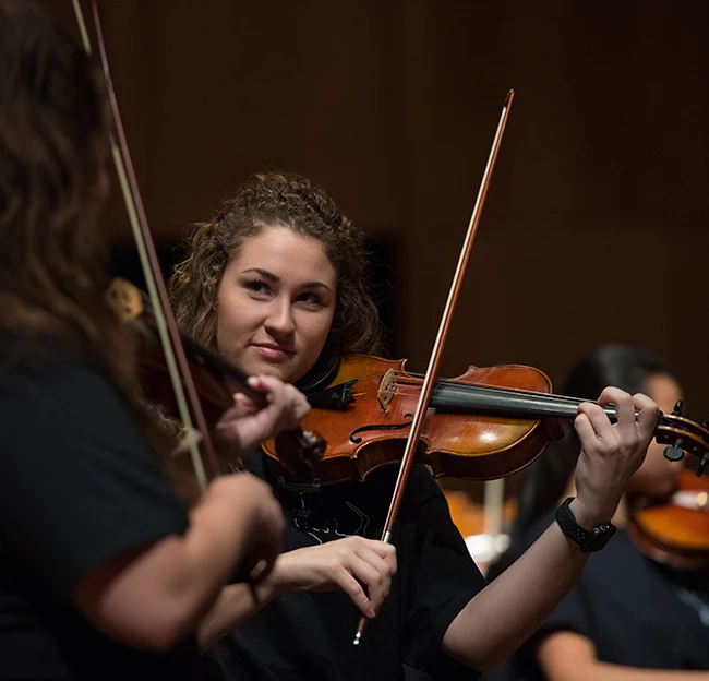 image of a female student playing the violin and smiling at another student