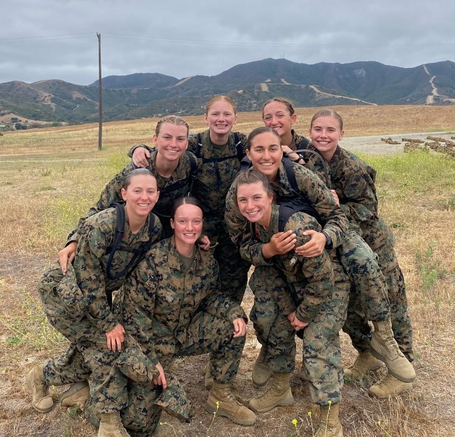 small group of female midshipmen smiling for the camera while standing outside in a field with mountains in the background