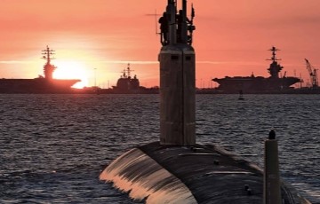 sunset over the water with a submarine in the foreground and aircraft carriers in background