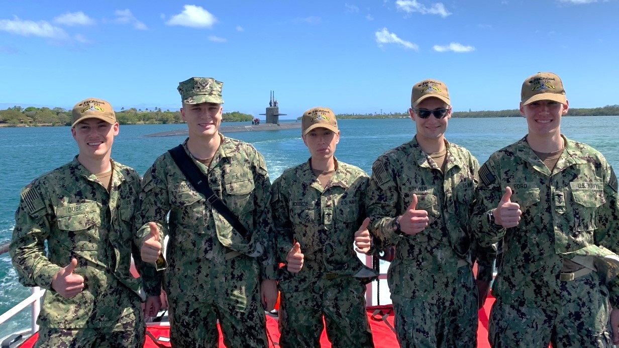 image of a small group of Midshipmen standing and smiling at the camera with a submarine in the water behind them