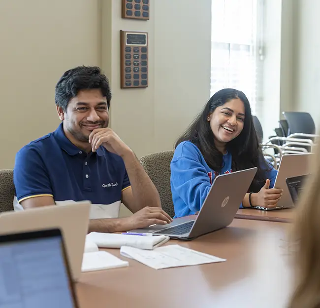 Two students sitting at a table behind open laptops smiling.