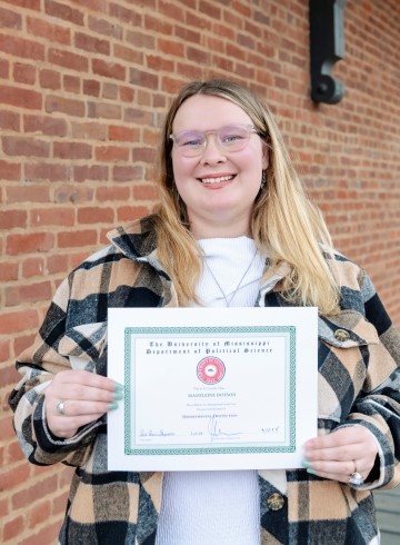 image of Madeleine Dotson standing in front of a brick wall smiling at the camera and holding up a certificate