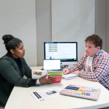 writing center mentor and a student seated in a Writing Center meeting room and looking at a writing handbook