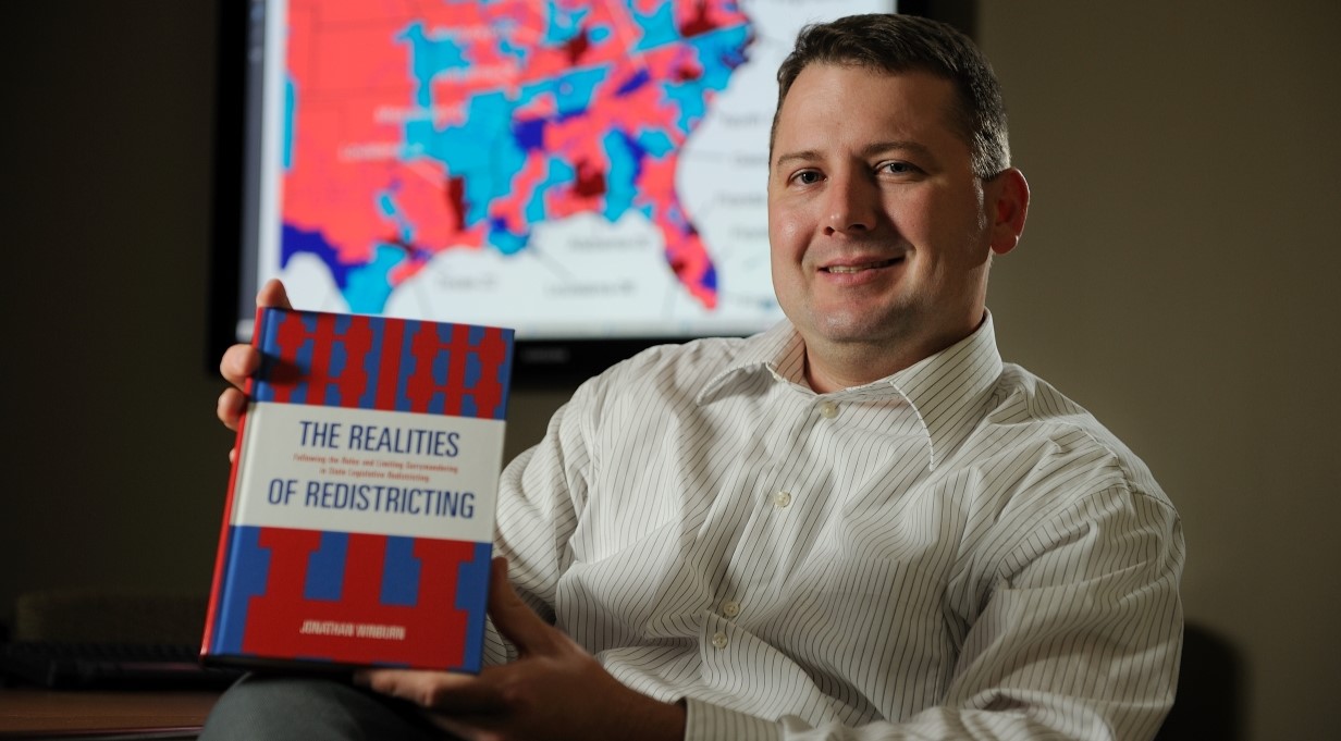 professor holding his book while seated in a chair in his office. Map of electoral districts on his computer screen in the background