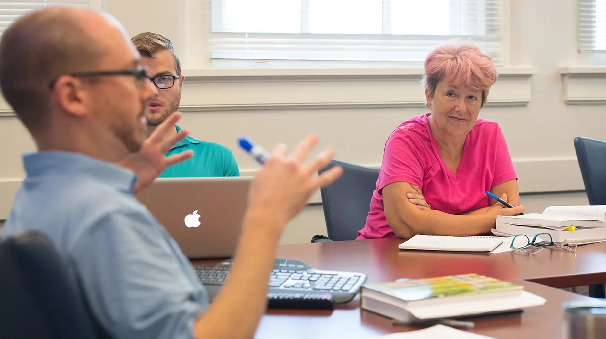 Professor in foreground and out of focus is seated at a table, hands gesticulating and holding a marker. Two students, in focus, are also seated at the table looking at professor.