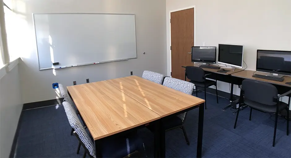 Room with blue carpet, large table with four chairs, tables and chairs with computers, and large white board.