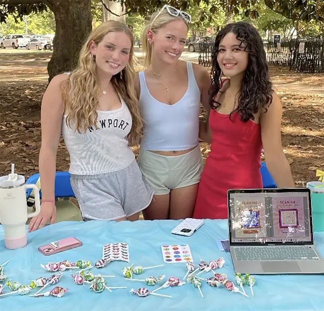 Three students, smiling and looking at the camera, standing behind table strewn with candy and a laptop.