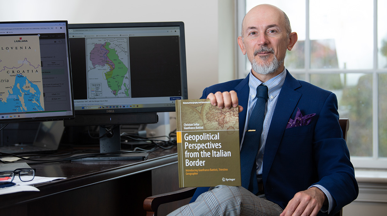 man sitting at desk holding his book with maps up on a computer screen in the background