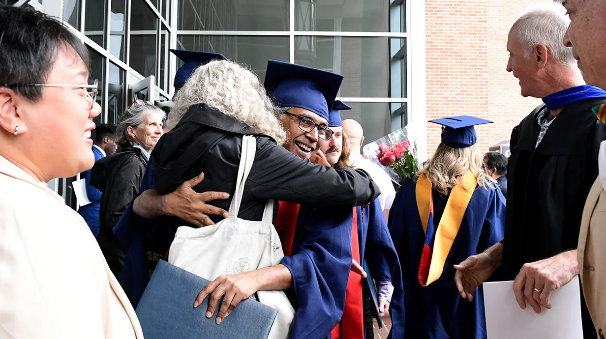 Scene from graduation: student in graduation robe hugging a person; other people look on smiling.. 