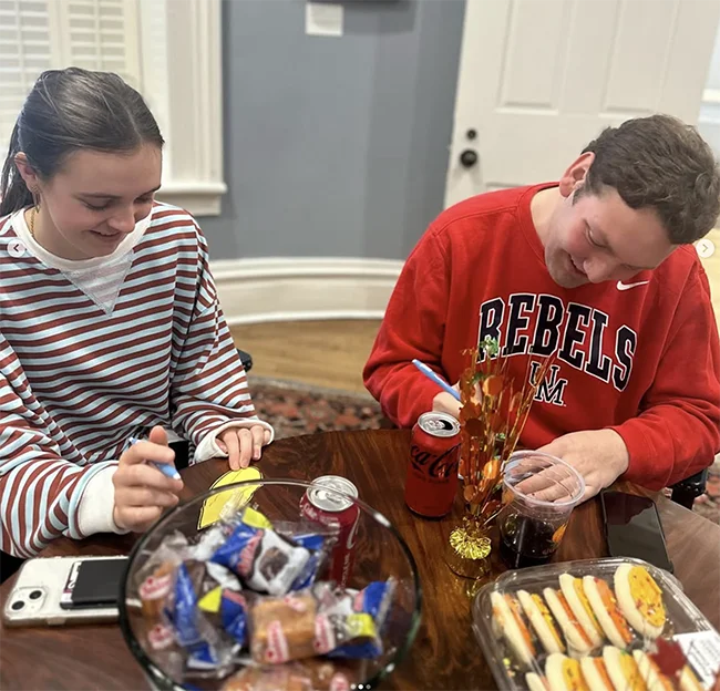 two people sitting at a table filled with snacks writing on paper and smiling