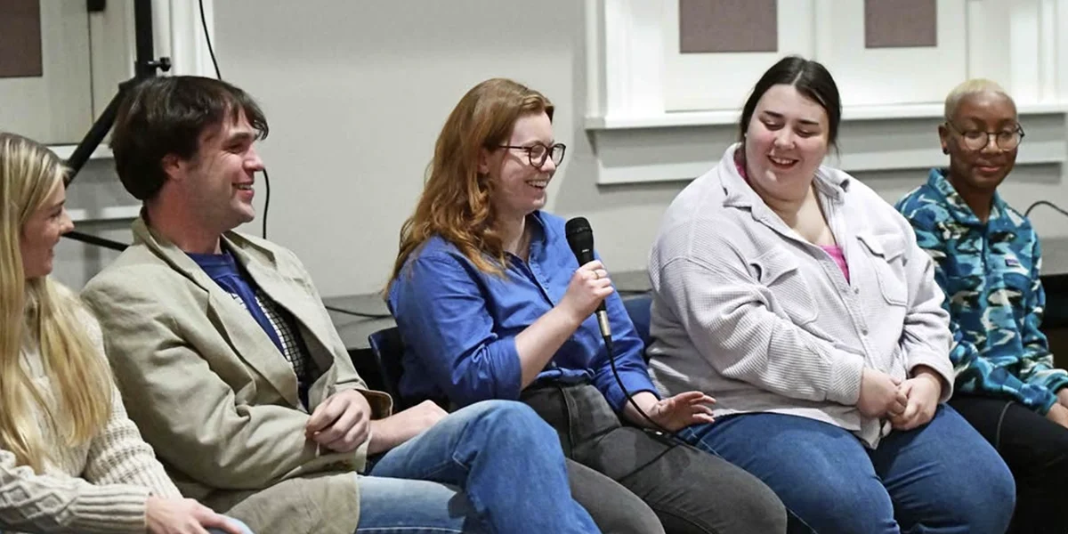 students sit in chairs on stage; one student holds a microphone