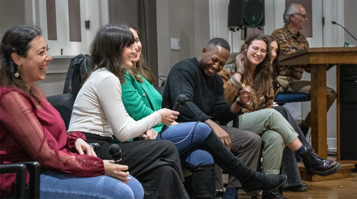 group of seven smiling and laughing people sit on a stage. Two people hold microphones