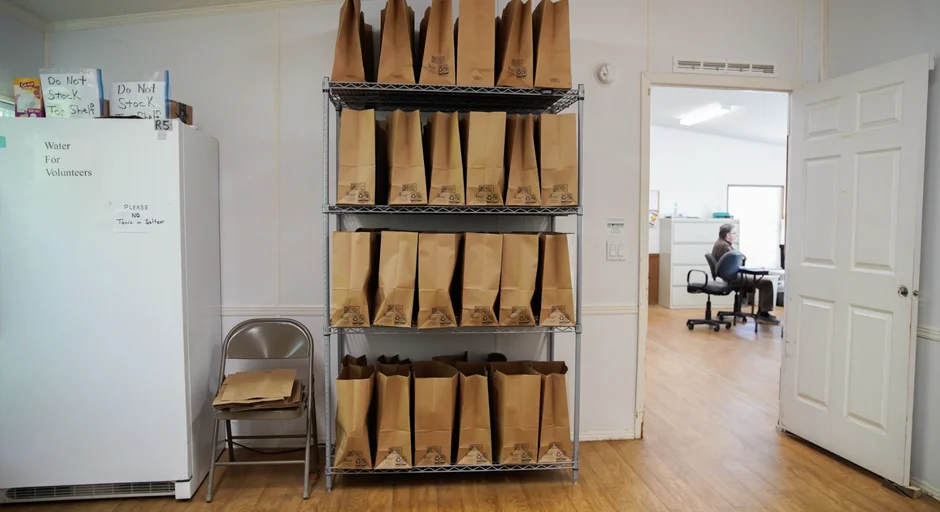 Room with refrigerator on left, shelves in the middle stacked with filled paper bags, and a door opened to an office on the right.