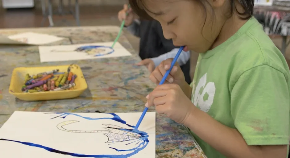 Child blows on blue paint on paper through a straw  