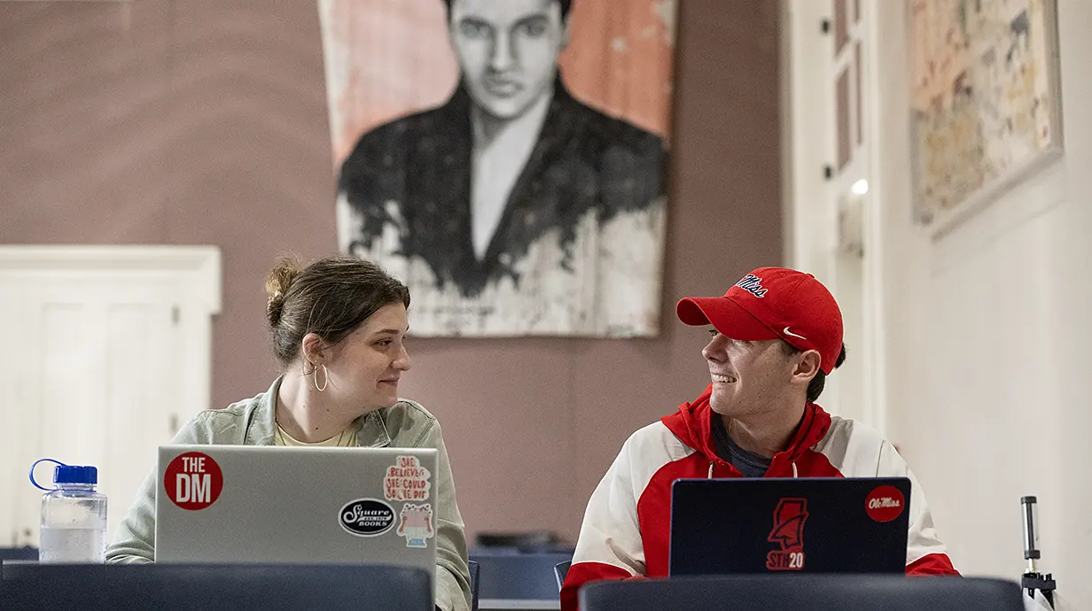 Two students behind laptops look at each other; large tapestry of Elvis hanging in background