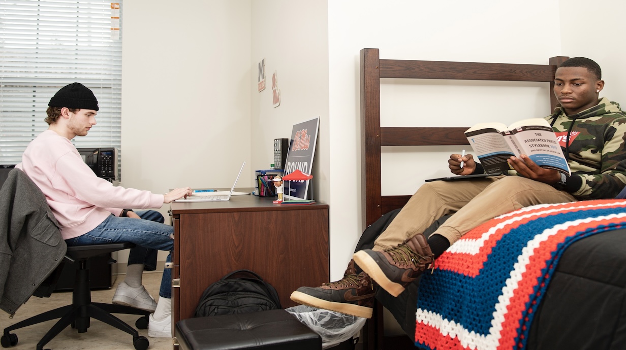 one student sitting at desk and another student sitting on a bed. 