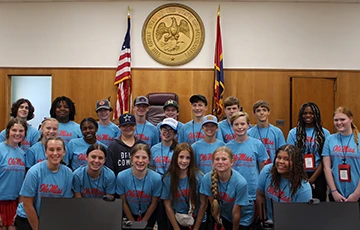 A group of high school students wearing matching blue Ole Miss T-shirts stand in a courtroom in front of the great seal of Mississippi.