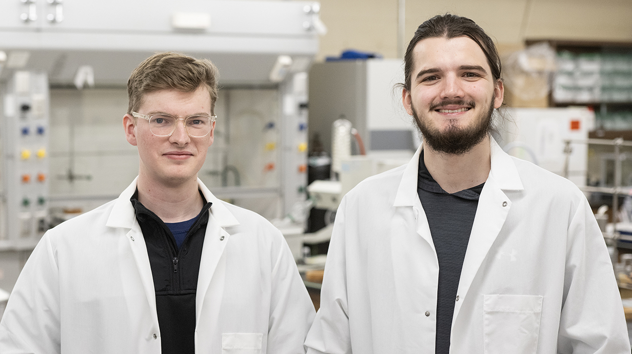 Two young men wearing white lab coats stand in a laboratory.