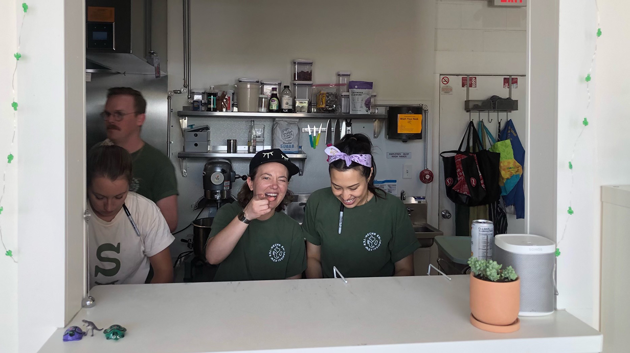 Three women and a man work in a kitchen behind a pass-through window.