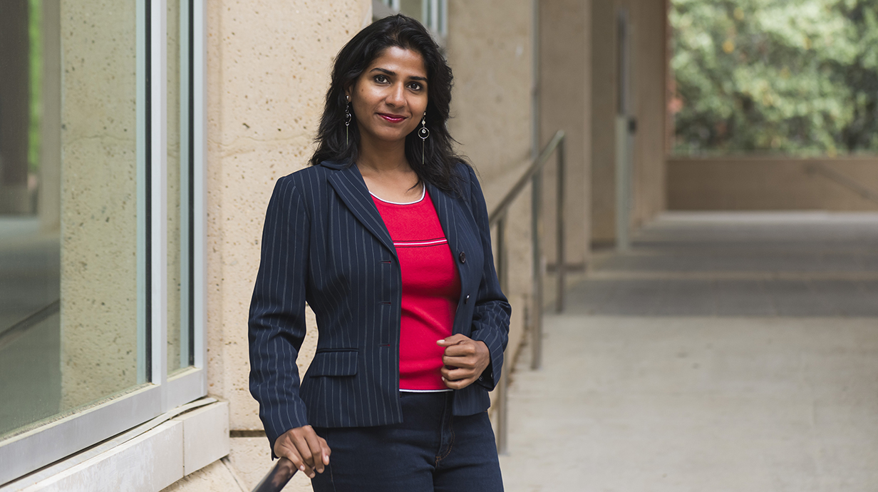 A woman wearing a blue blazer and a red top leans against a railing outside a building.