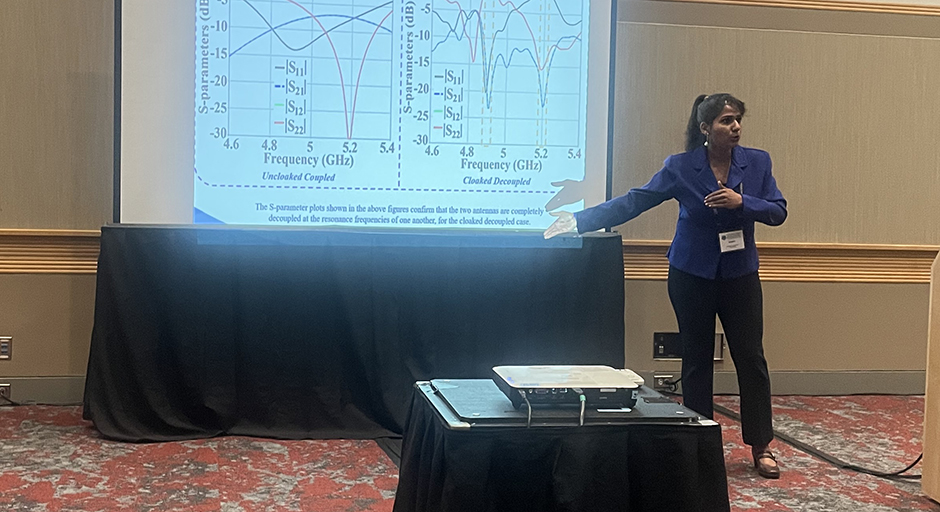 A woman stands in a conference room, pointing at a screen displaying engineering diagrams.